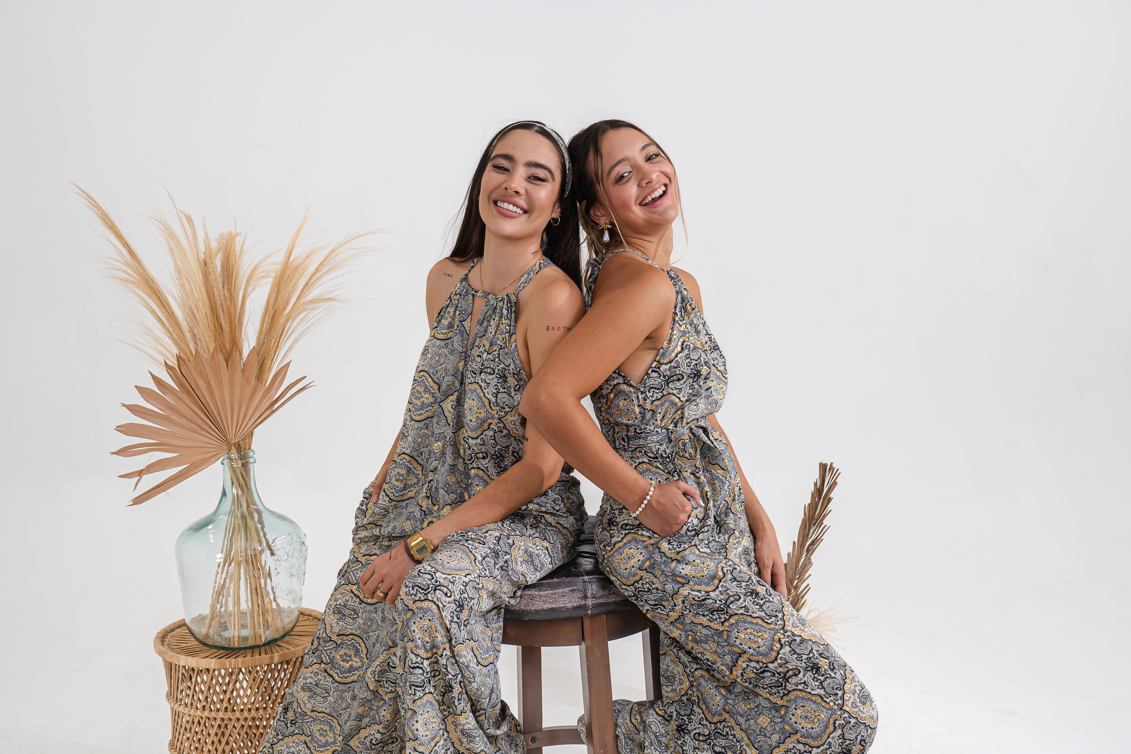 Two women in matching patterned dresses sitting together with a plant on a white background